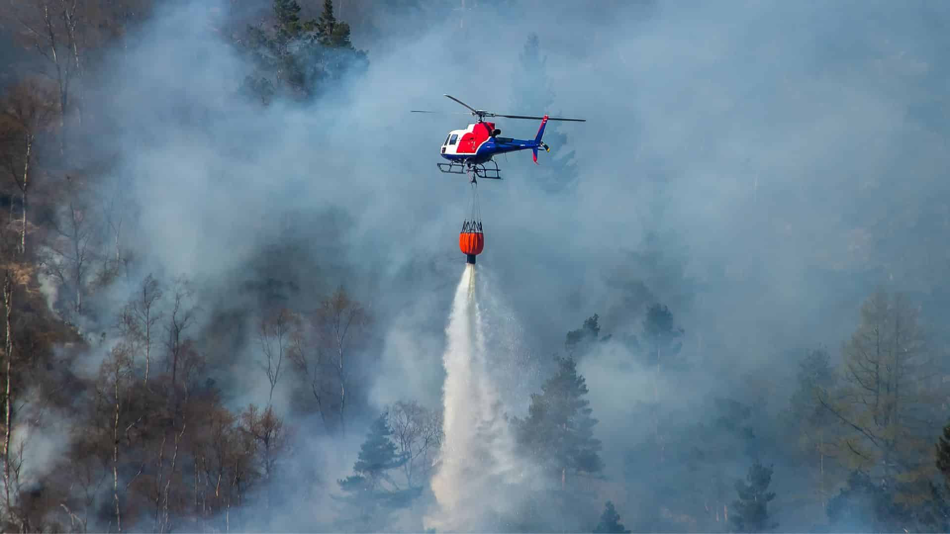 A helicopter extinguishes a forest fire as smoke drifts across the area.